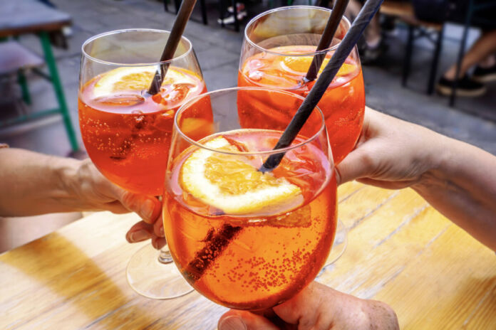 Drei Gäste prosten sich mit Aperol Spritz in großen Weingläsern zu. In jedem Glas befinden sich Prosecco, Aperol, eine Orangenscheibe und Eis. Außerdem haben sie noch einen schwarzen Strohhalm bekommen. Sie sitzen in einem Biergarten auf einer großen Terrasse.