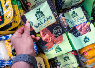 Die Hand eines Kunden greift in das Kühlregal eines Supermarktes oder Discounters und nimmt eine Packung veganer Salami heraus. Neben diesem Produkt befinden sich noch viele weitere vegane Produkte im Kühlregal.