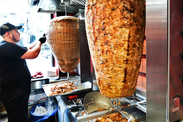 In einem Dönerladen drehen sich saftige Dönerspieße. Ein Mitarbeiter schneidet Fleisch von einem Dönerspieß ab, um es in einem leckeren Döner zu servieren und an seine Kunden auf der Straße zu verkaufen.