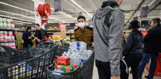 In einem Discounter oder Supermarkt haben einige Kunden ihre Einkaufswagen bis oben hin mit Getränken vollgepackt. Im Hintergrund schauen sich Kunden einige Artikel an. Der Markt ist gut besucht.