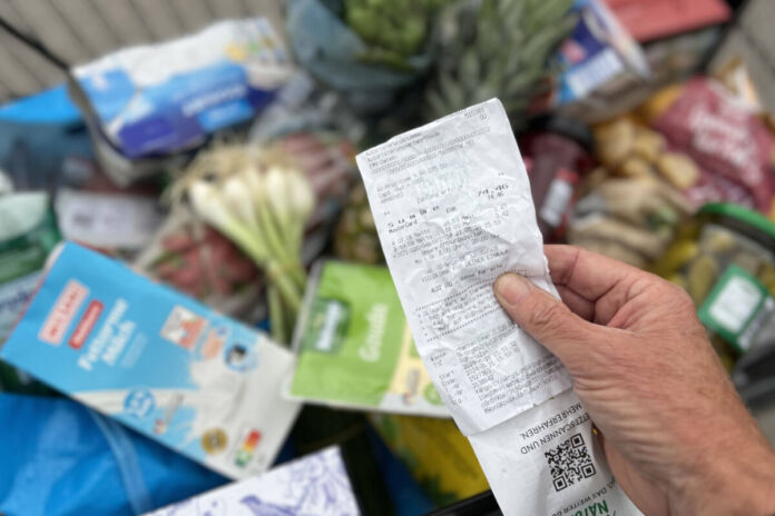 Eine Person hält einen Einkaufszettel in der Hand und überprüft ihn. Im Hintergrund befindet sich ein voller Einkaufswagen mit Grundnahrungsmitteln. Um das Leben finanzieren zu können, müssen Bürger ungefähr 4.000 Euro verdienen.