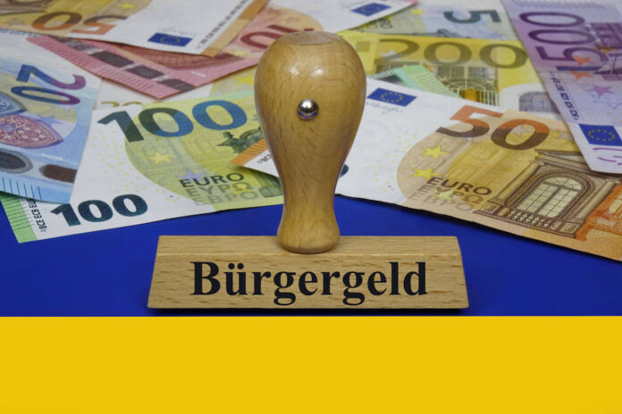 Ein Stempel mit dem Aufdruck "Bürgergeld" liegt auf einer ukrainischen Flagge. Hier liegt auch eine Menge Bargeld, das staatliche Zuschüsse symbolisiert. Es sind 5 Euro, 10 Euro, 20 Euro, 50 Euro, 100 Euro, 200 Euro und ein 500-Euro-Schein.