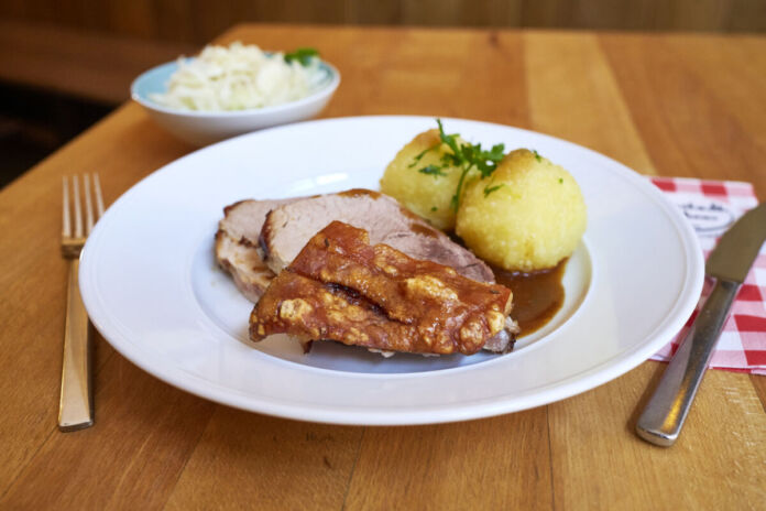 Ein Essen im Restaurant. Es handelt sich um ein typisch deutsches oder bayerisches Gericht. Es besteht aus einem Schweinebraten mit Kruste und Kartoffelknödeln. Rechts und links liegen Gabel und Messer. Im Hintergrund lässt sich ein Beilagensalat erkennen.