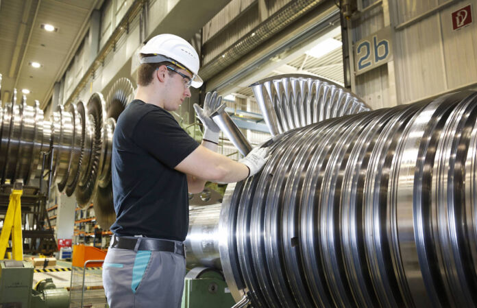 Ein Facharbeiter in der Fabrik bei der Arbeit. Er trägt Schutzbrille und Schutzhelm und hantiert an einer großen Maschine aus Stahl oder Metall. Er ist Angestellter eines Weltkonzerns aus Baden-Württemberg.