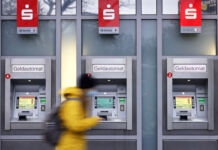 Ein Fußgänger mit einer gelben Jacke geht an drei Geldautomaten der Sparkasse vorbei. Unter dem Logo der Sparkasse steht jeweils die Bezeichnung SB-Service.