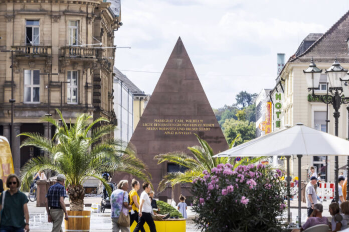 Die Pyramide steht als Wahrzeichen auf dem Karlsruher Marktplatz. Bürger laufen durch die Innenstadt und bummeln durch die Läden und Gaststätten.