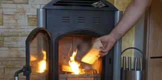 Ein Mann legt Feuerholz im Kaminofen nach, der den Raum beheizt. Die Kamintür und das Gitter sind geöffnet. Im Ofen brennen schon ein paar Scheite, die eine behagliche Stimmung schaffen.
