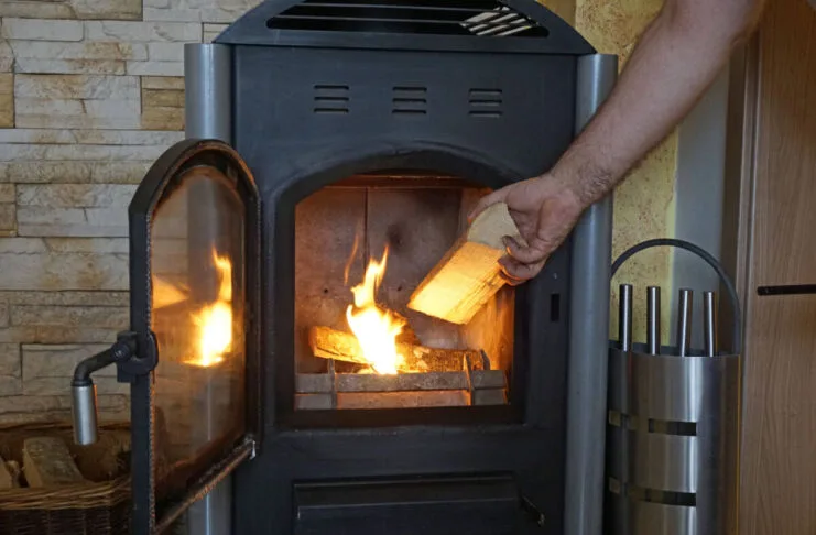 Holzofen Kamin Wohnzimmer Feuer Holz Brennholz beheizen befeuern Hand Kamingitter Kamintür Holzscheit Ein Mann legt Feuerholz im Kaminofen nach, der den Raum beheizt. Die Kamintür und das Gitter sind geöffnet. Im Ofen brennen schon ein paar Scheite, die eine behagliche Stimmung schaffen.