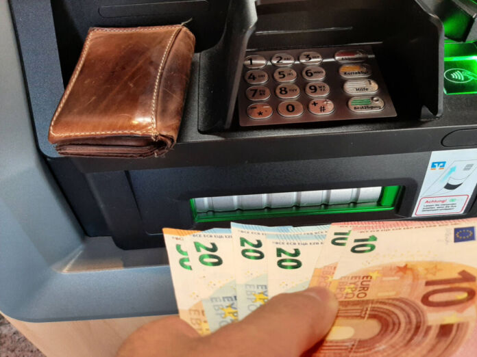 Ein Mann oder eine Frau steht an einem Geldautomaten und hebt Geld ab. In der Hand hält die Person mehrere Geldscheine. An dem Gerät liegt auch das Portemonnaie.