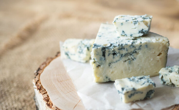 Auf einer Holzplatte vor einem hellen Hintergrund liegt ein schönes Stück Käse. Es handelt sich um Schimmelkäse, deutlich an den grün-bläulichen Flecken zu erkennen.