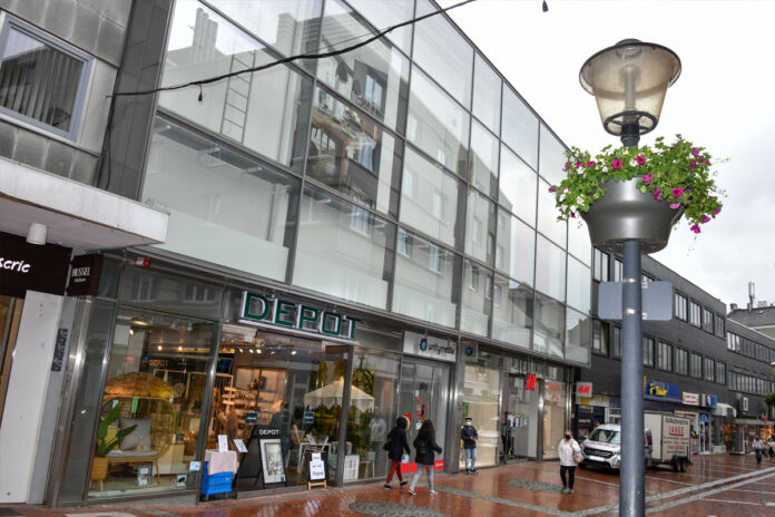 Ein Blick auf eine Filiale der bekannten Deko-Kette Depot. Das Geschäft befindet sich in einer Einkaufsstraße und wurde offenbar an einem regnerischen Tag fotografiert. Einige Menschen gehen daran vorbei. Sie tragen dicke Jacken. Der Asphalt ist nass.