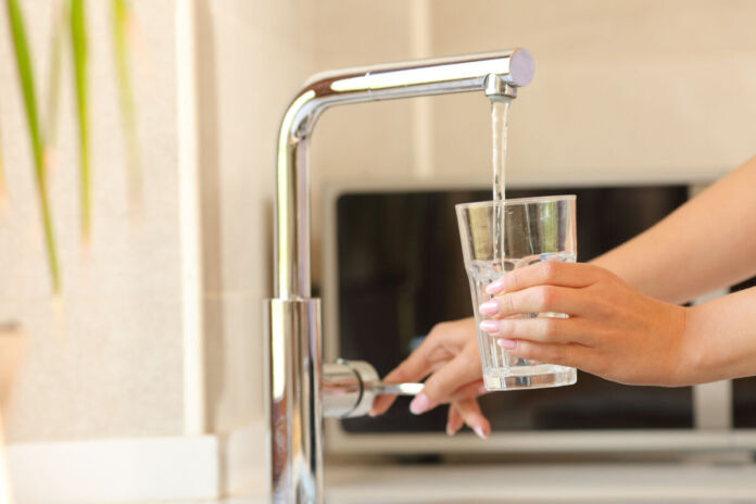 Eine Frau hält ein Glas unter einen Wasserhahn und lässt Leitungswasser in das Trinkglas laufen. Im Hintergrund sieht man verschwommen die Küche. Am Fenster steht eine Pflanze.