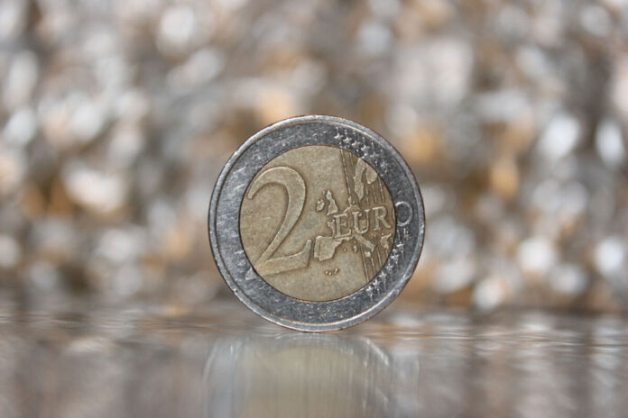 Auf einem Tisch mit einer spiegelnden Oberfläche steht eine 2-Euro-Münze. Das Bild zeigt eine Nahaufnahme der Münze. Der Hintergrund ist verschwommen und man kann keine konkreten Elemente sehen.