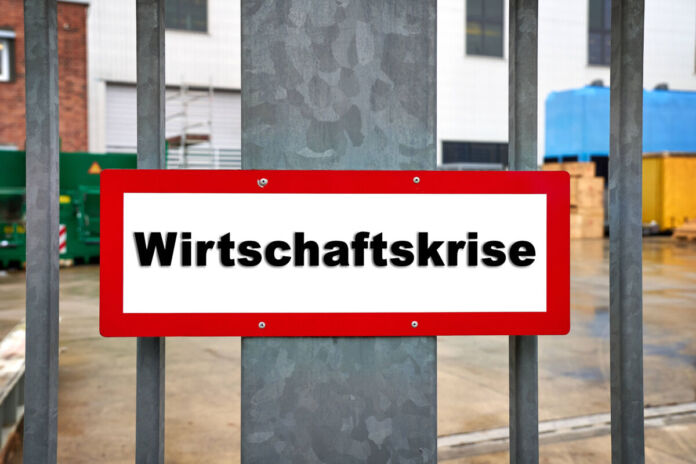 Ein weißes Schild mit schwarzer Beschriftung und einer roten Umrandung. Die Inschrift lautet "Wirtschaftskrise". Das rechteckige Schild ist in einem Industriegebiet an einem grauen Tor befestigt.