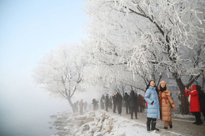 Es ist tiefster Winter und auf einem Weg am Wasser ist alles eingeschneit. Die Bäume sind mit Frost benetzt. Der Himmel ist strahlend blau. Auf dem Weg laufen Menschen in dicken Mänteln, und zwei Frauen in Daunenmänteln lassen sich vor der winterlichen Landschaft fotografieren.