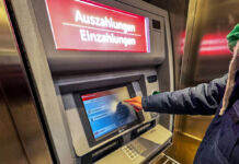 Eine unbekannte Person steht vor dem Geldautomaten einer Bank und wählt gerade aus dem Bildschirmmenü per Touch-Funktion die gewünschte Funktion aus.