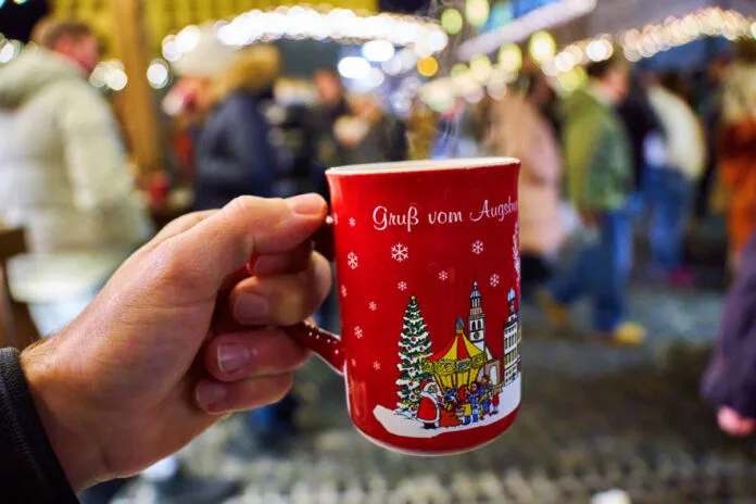 Weihnachten Weihnachtsmarkt Weihnachtsbude Weihnachtsstand Glühwein Tasse Getränk Stiefel Besucher Die Hand eines nicht erkennbaren Mannes hält eine Tasse Glühwein in die Kamera. Die Tasse ist ein typischer Becher für einen Weihnachtsmarkt. Im Hintergrund sieht man viele Menschen, die den Weihnachtsmarkt besuchen und an den verschiedenen Ständen stehen.