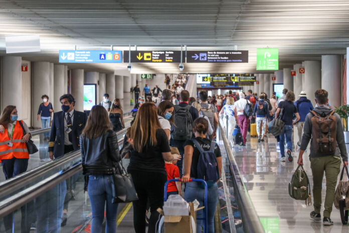 In einem Flughafen sind viele Menschen unterwegs, die einen langen Flur zu ihrem Terminal entlanggehen. Mit sich führen sie Koffer und Reisegepäck.