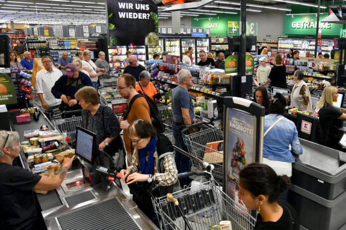 In einer großen Kaufland-Filiale tummeln sich viele Menschen, die ihre Lebensmittel einkaufen. Einige warten an der Kasse, bis sie an der Reihe mit dem Bezahlen sind, während ein Kassenmitarbeiter die Produkte scannt.
