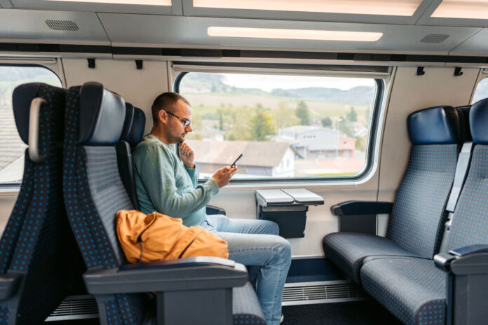 An einem Viererplatz in einem gemütlichen Schnellzug sitzt ein Mann und schaut auf sein Handy. Im Fenster neben ihm sieht man die Landschaft und auf seinen Nebenplatz hat der Zugpassagier seine Tasche gelegt.