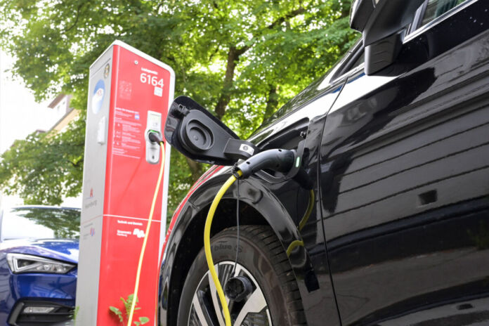 Ein Elektroauto wird aufgeladen. Es befindet sich an einer öffentlichen Ladesäule. Das Stromkabel ist eingesteckt. Im Hintergrund kann man erkennen, dass das Auto in einer Straße steht und die Bäume ein sommerliches Grün tragen.
