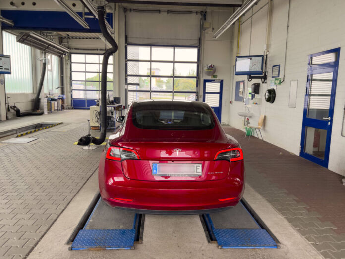 Ein roter Tesla steht auf zwei Schienen in einer Werkstatt. Das Elektroauto soll wohl repariert werden oder die Angestellten sollen eine Hauptuntersuchung vornehmen.