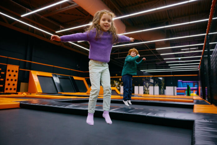 Zwei Kinder, ein Junge und ein Mädchen, springen auf einem Trampolin in einer großen Halle, die zu einem Indoor-Spielplatz gehört.