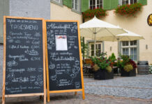 Auf den Steinen einer Fußgängerzone stehen zwei große Tafelschilder vor einem Restaurant. Auf diesen finden sich zahlreiche Gerichte, die das Lokal anbietet, sowie die Preise dafür.