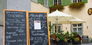 Auf den Steinen einer Fußgängerzone stehen zwei große Tafelschilder vor einem Restaurant. Auf diesen finden sich zahlreiche Gerichte, die das Lokal anbietet, sowie die Preise dafür.