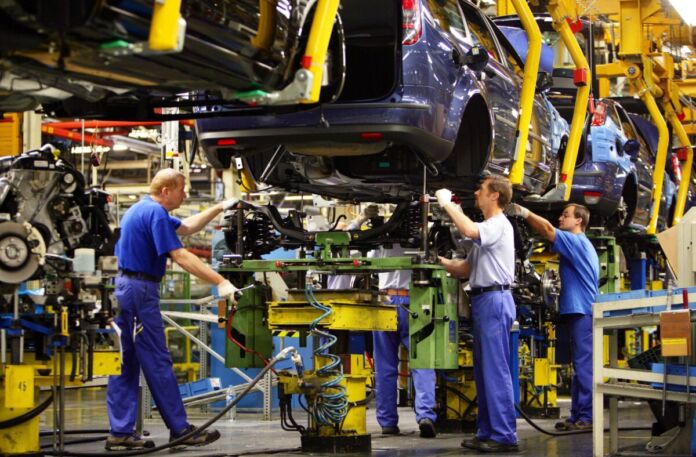 In einer großen Werkhalle mit Maschinen bauen mehrere Arbeiter einen Motor in die Karosserie eines Autos der Marke Ford ein. Dieser Prozess wird auch "Hochzeit" in der Autoherstellung genannt.