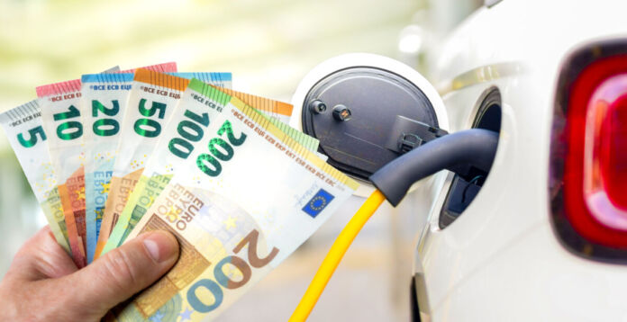 Eine männliche Hand hält mehrere Euro-Scheine in die Kamera. Daneben befindet sich ein E-Auto, das gerade an eine Ladestation angeschlossen zu sein scheint. Elektroautos werden jetzt immer günstiger.