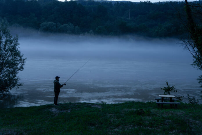 Ein Mann steht an einem Fluss, welcher durch das Gebirge fließt. Der Angler versucht noch in der Nacht Fische zu fangen. Das Wasser ist kaum zu sehen, weil es dunkel ist.