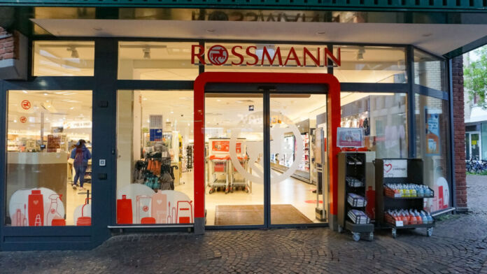 Der Blick fällt in eine geöffnete Rossmann-Filiale, die beleuchtet ist. Draußen vor dem Eingang stehen in mehreren Regalen verschiedene Produkte, die die Drogerie anbietet. Im Inneren der Filiale laufen Kunden durch das Geschäft.