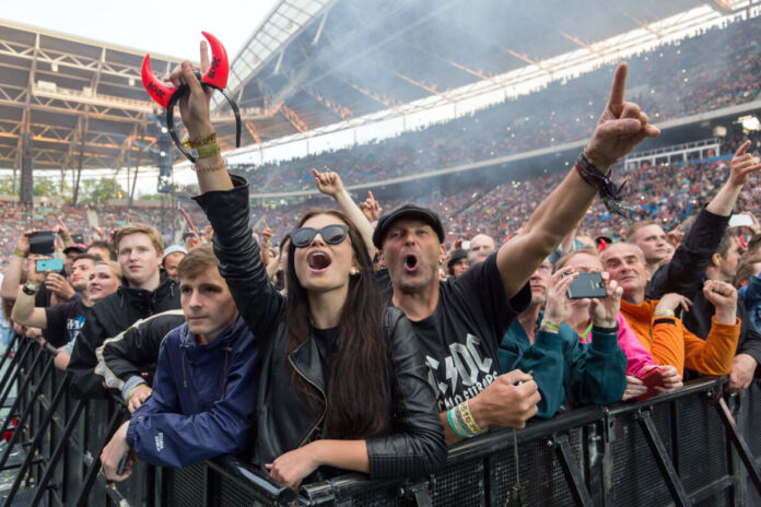 Eine große Menschenmenge hat sich in einer Arena zu einem AC/DC-Konzert versammelt. Die Zuschauer tragen Kleidung mit dem Logo der Band und jubeln dem Open Air zu. Vor der Bühne stehen Tausende.