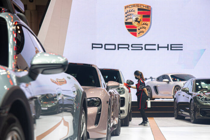 Auf einer großen beleuchteten Fläche stehen mehrere neue Fahrzeuge von Porsche in einer Reihe. Eine Mitarbeiterin hat ein Tuch in der Hand und poliert offenbar nach. Im Hintergrund sieht man vor einer großen hellen Wand das Porsche-Logo und darunter den Namen Porsche in Großbuchstaben.