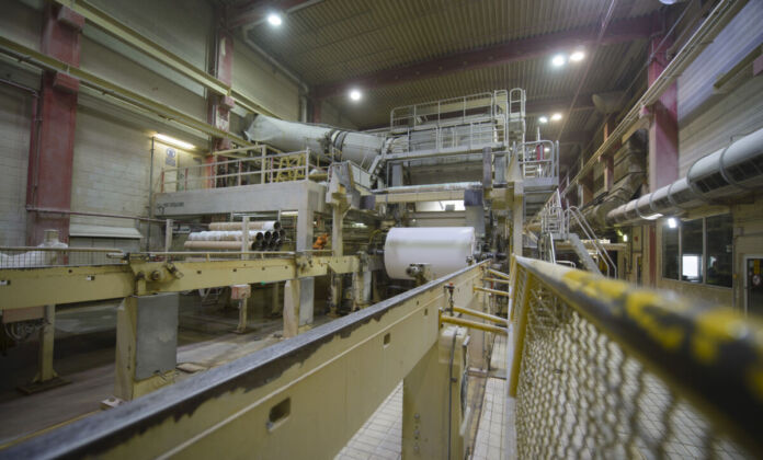 Industrielle Anlage eines Papierherstellers in einer Halle. Auf einer Ablage befinden sich mehrere Papierrollen. Viele Teile der Anlage sind gelb, einige grau. Die Halle ist beleuchtet und menschenleer.