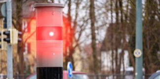 Im Vordergrund steht eine rot leuchtende Blitzersäule und kontrolliert den Verkehr auf Temposünder. Die Verkehrsüberwachung dient der Sicherheit auf den Straßen für Autos und Fußgänger. Im Hintergrund ist eine Straße mit Autos zu sehen.