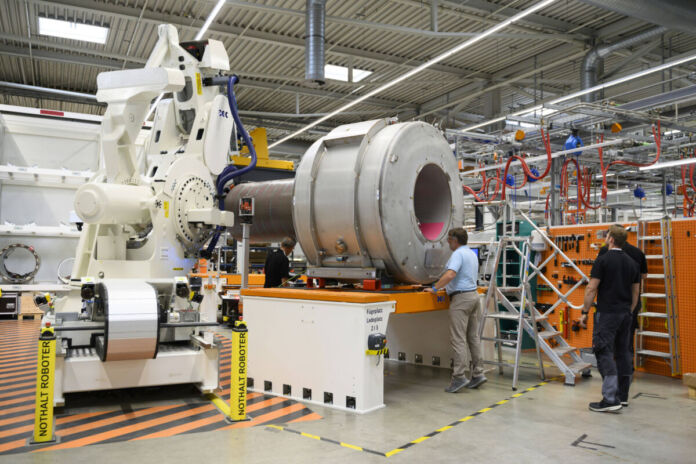 Eine Produktionshalle für Magnetresonanztomographen. In der Halle sind vier Menschen und verschiedene Geräte, Leiter und Werkzeuge zu sehen. Ein weißer Roboter, bei dem es sich um einen "Nothalt-Roboter" handelt, ist in der Halle.