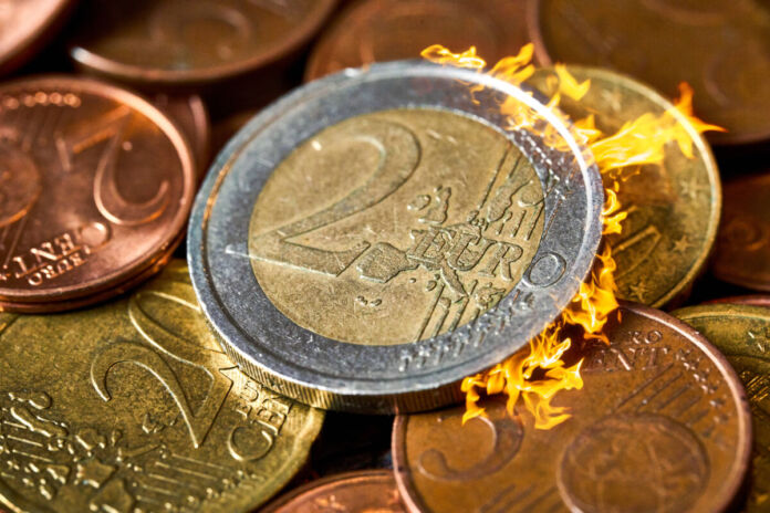 Auf einem Haufen voller 2-, 5- und 20-Cent-Stücke, die kreuz und quer durcheinander liegen, befindet sich ein 2-Euro-Stück. Auf der rechten Seite der Münzen schlagen Flammen heraus.