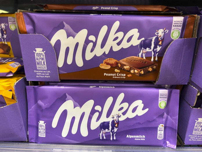 In einem Regal in einem Supermarkt oder Discounter sind verschiedene Sorten von Schokolade der bekannten Marke Milka einsortiert. Die Tafeln befinden sich in Kartons und sind übereinander gestapelt. Die Traditions-Schokolade Milka ist jetzt fast doppelt so teuer.