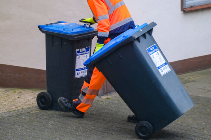 Ein Mitarbeiter der Abfallentsorgung mit zwei Mülltonnen. Bei den Mülltonnen handelt es sich um die schwarze Papiertonne mit blauem Deckel. Der Mitarbeiter schiebt eine Tonne zurück an ihren Platz, während er zur anderen Tonne greift.