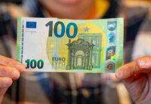 Eine Person, die sich im Hintergrund befindet und nur verschwommen von der Kamera erfasst wird, hält einen 100-Euro-Schein mit beiden Händen in die Kamera. Der Euro-Schein sieht noch sehr neu aus, da er sauber und nicht zerknittert ist.