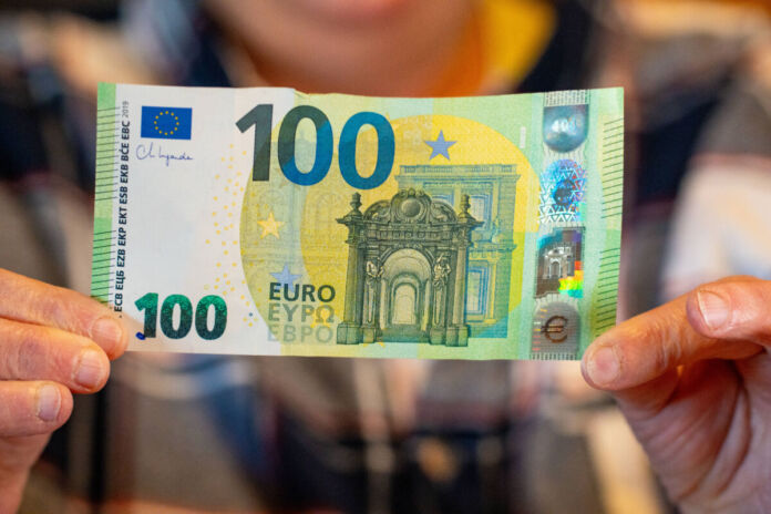 Eine Person, die sich im Hintergrund befindet und nur verschwommen von der Kamera erfasst wird, hält einen 100-Euro-Schein mit beiden Händen in die Kamera. Der Euro-Schein sieht noch sehr neu aus, da er sauber und nicht zerknittert ist.