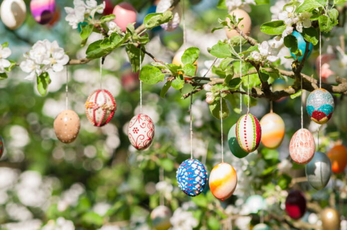Mehrere bunte Ostereier, die als Dekoration am Ast eines Baumes hängen. Es sind weiße Blüten und grüne Blätter an dem Ast zu sehen, die auf den Frühling hinweisen.