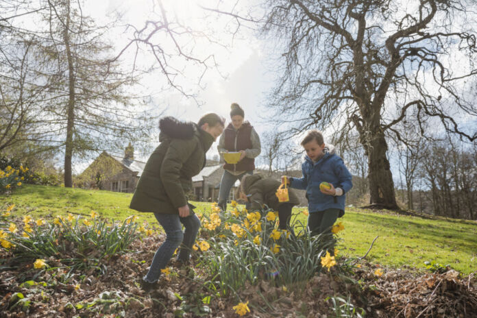 Zwei Erwachsene und zwei Kinder draußen im Garten. Es ist Frühling und die Sonne scheint. Die Narzissen blühen und gemeinsam werden Ostereier gesucht. Es scheint Ostersonntag zu sein und die Kinder tragen kleine Osterkörbe.