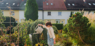 Eine junge Frau im Jeans-Overall steht in ihrem Garten. Die Frau hält eine Gießkanne mit Wasser und begießt eine Pflanze. Im Garten befinden sich verschiedene Pflanzen und Bäume.
