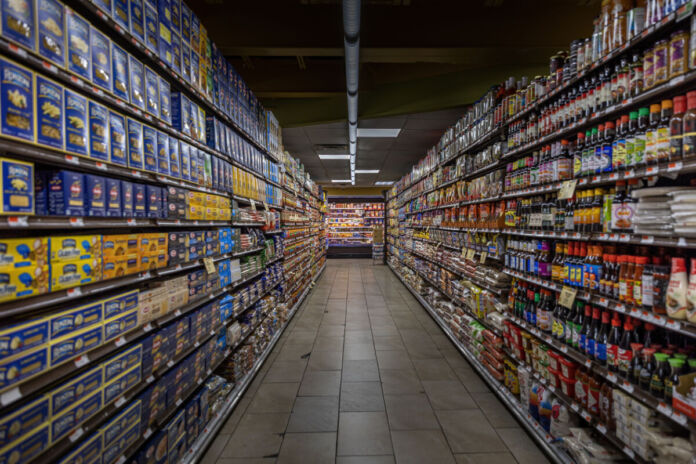 Auf dem Bild ist ein Supermarktgang zu sehen. Links und rechts stehen hohe Regale mit zahlreichen Produkten. Auf der linken Seite befinden sich überwiegend Packungen mit Pasta in blauen und gelben Verpackungen. Auf der rechten Seite sind verschiedene Lebensmittel wie Soßen, Gewürze und Reis zu sehen.
