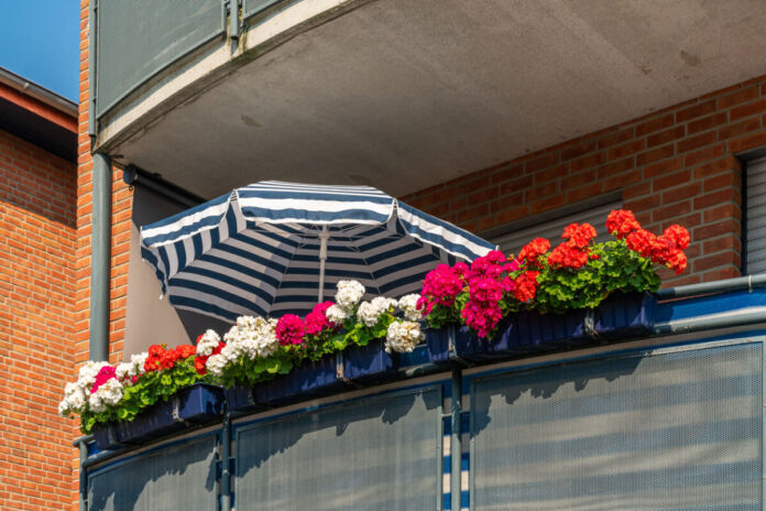 Der Blick fällt auf den Sichtschutz eines Balkons einer Wohnung. In den blauen Blumenkästen befinden sich verschiedene bunte Blumen. Ein Sonnenschirm wurde aufgespannt. Die Jalousien sind heruntergelassen.