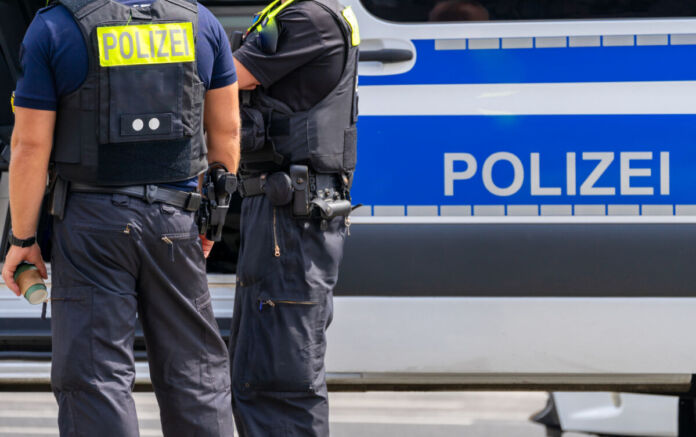 Zwei Beamte von der Polizei stehen in Uniform vor einem Einsatzwagen. Die Behörden sichern den Tatort. In Karlsruhe kam es zu einem brutalen Raubüberfall.