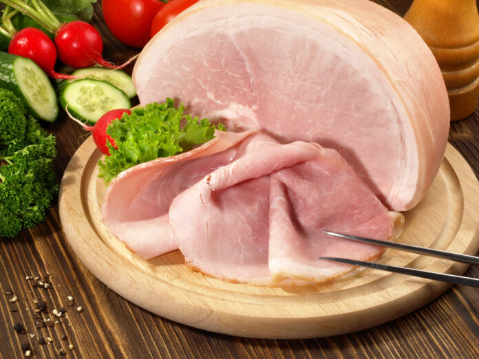 Auf einem Brett aus Holz liegt ein Stück Schinken, von dem eine dünne Scheibe abgeschnitten wurde. Im Hintergrund liegt Gemüse, darunter Gurken, Radieschen und Tomaten sowie ein paar Kräuter und Gewürze.